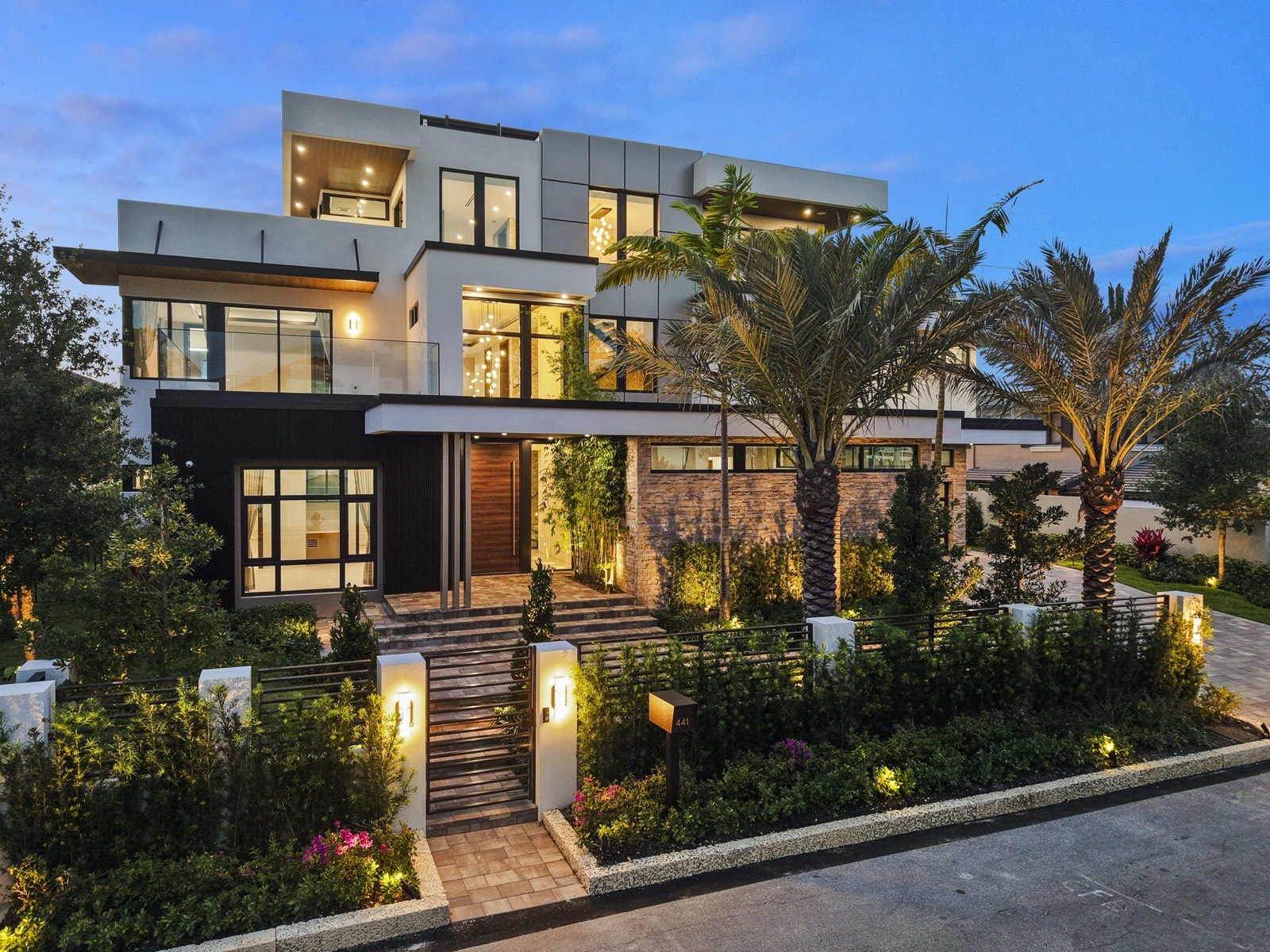 Three-story modern waterfront residence at dusk, seen from the street through palms